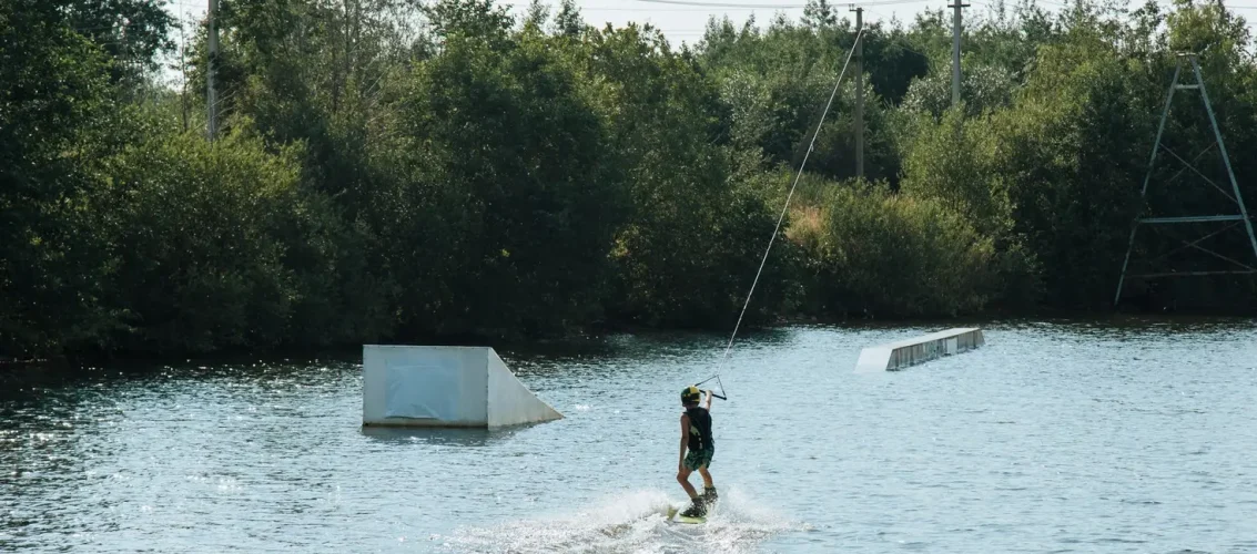 Praticante iniciante segurando uma prancha de wakeboard na beira do cable park em dia ensolarado