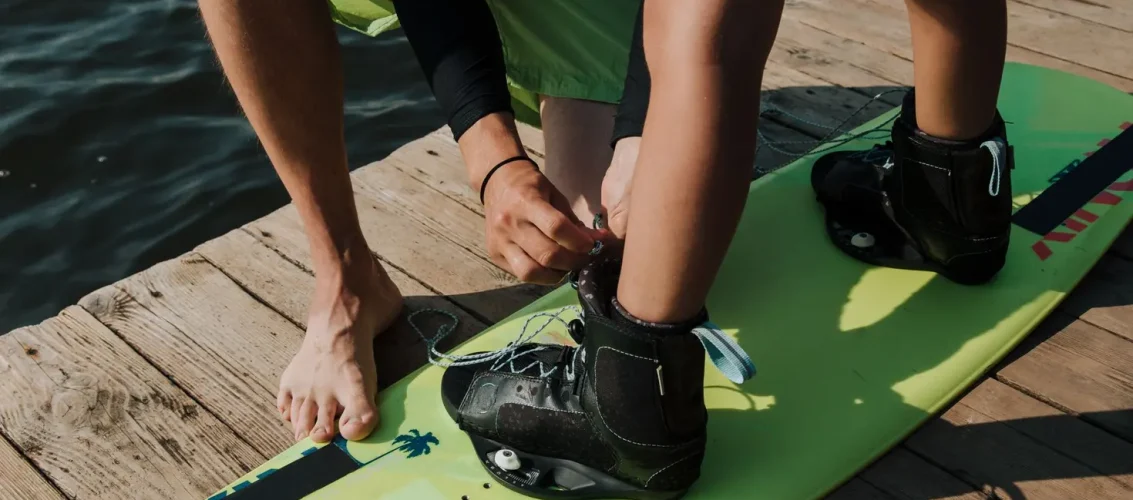 Pessoa inspecionando prancha de wakeboard usada na beira do lago, verificando base e inserts