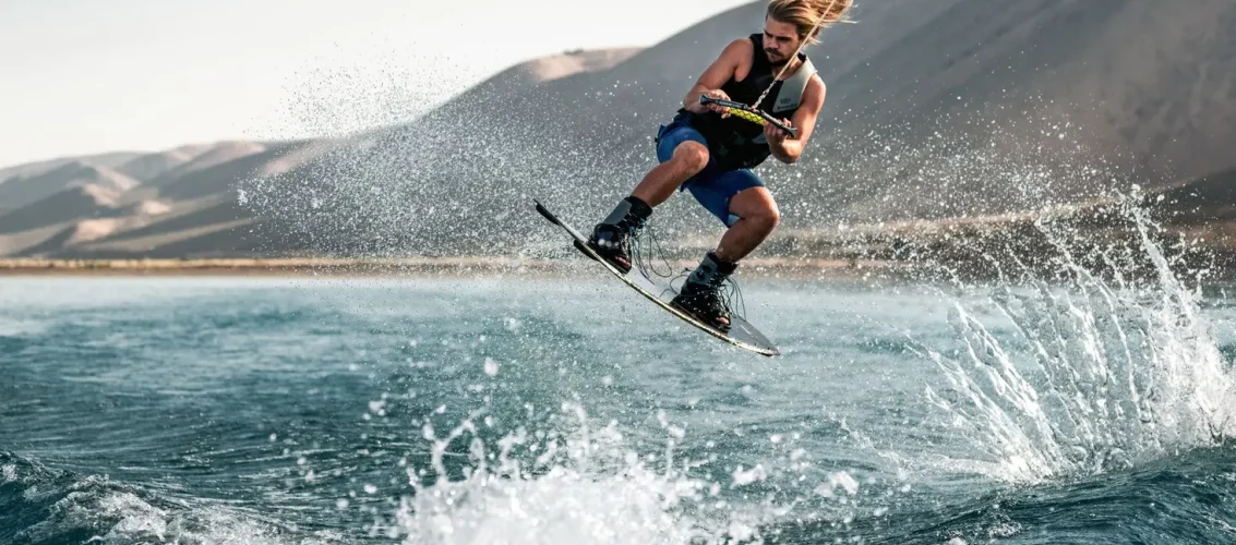 Praticante de wakeboard saltando na wake com prancha durante sessão em lago ensolarado, ilustrando os tipos de prancha de wakeboard
